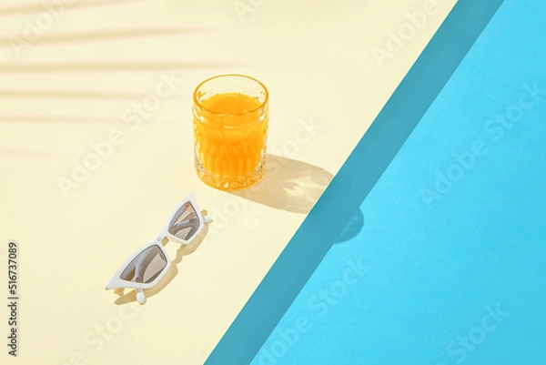 Obraz Minimal tropical composition with stylish white sunglasses and orange drink on duo tone sandy and blue background. Creative copy space with palm leaf shadow. Pool party concept.