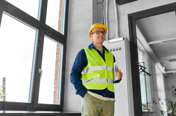 Fototapeta construction business and building concept - happy smiling male electrician or worker in helmet and safety west with papers on clipboard and pencil at electric board