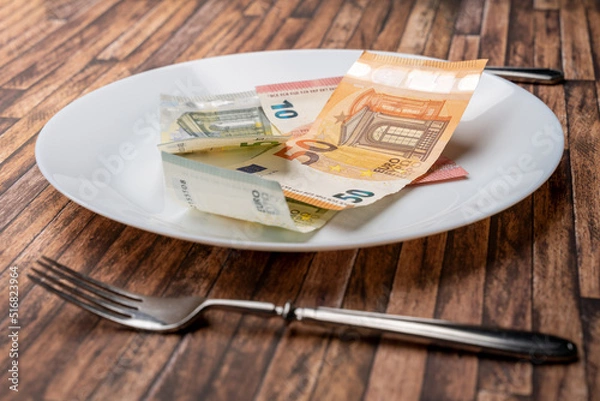 Obraz Euro banknotes lying on a dinner plate with cutlery on the table. Symbolic image for eating money notes. Food is getting more expensive due to inflation effects. Hunger caused by a crisis.