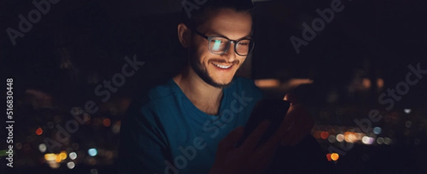 Obraz Night portrait of smiling man using smartphone near window. Panoramic banner.