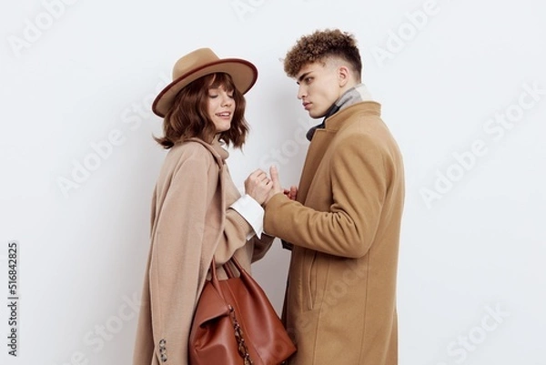 Fototapeta a happy couple in love is standing in beige stylish autumn coats on a white background and a man is holding a woman's hands smiling pleasantly