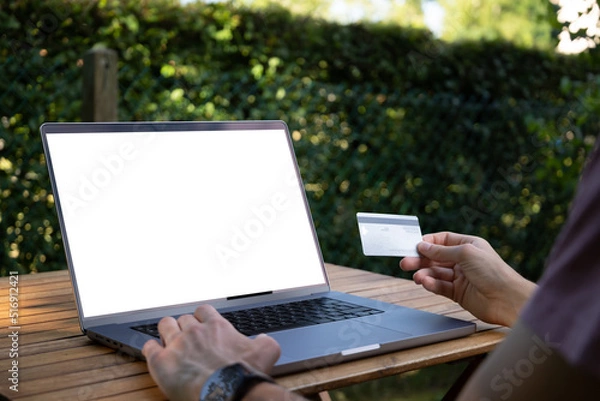 Obraz Jeune homme utilisant sa carte de crédit pour payer en ligne sur son ordinateur, vue rapprochée. Nomade numérique travaillant à l'extérieur, maquette écran d'ordinateur blanc