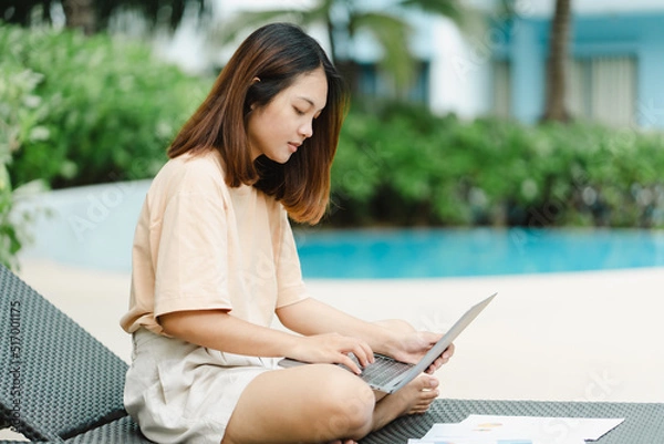Fototapeta A woman working at the swimming pool with her laptop computer holding paperwork in her hand. It is imperative for young Asian female tourists to sit and work on vacations at the hotel.