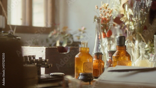 Fototapeta Close-up of a perfumer's workplace. Vintage cinematic concept. Sunny day in the working workshop. Lots of ingredients, a book, glass flasks on the table. Depth of field