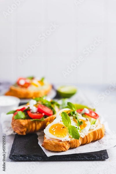 Obraz Variety of croissant sandwiches with tomato, cucumber, mozzarella cheese, eggs and mashed avocado with mint and arugula. Open savory sandwiches. Tasty healthy breakfast.