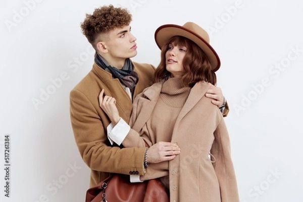 Fototapeta a sweet, joyful, pleasant stylish couple stands on a white background in autumn coats and gently hug each other, a man holds a woman by the coat looking at her