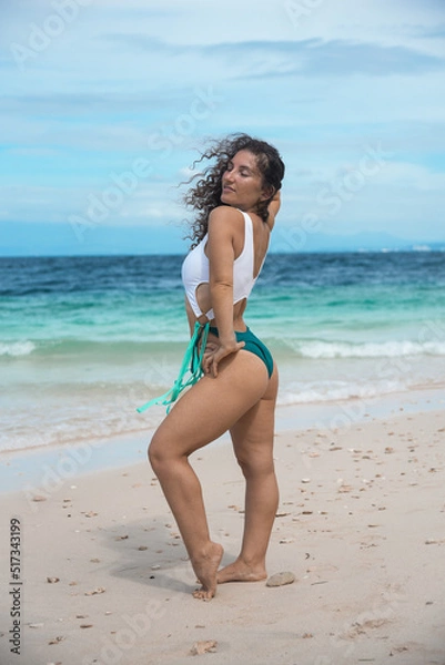 Fototapeta Beautiful female in a colorful bikini on a tropical beach. A brunette girl in swimsuit enjoy and walks on the beach. Happy young curly-haired woman smiling at the sea.