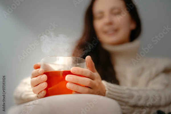 Fototapeta A young woman warms her hands on a glass mug with tea on a winter evening at home. The concept of the common cold.