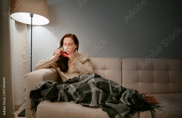 Fototapeta Young woman caught a cold by wrapping herself in a blanket at home and drinking hot tea from a cup.