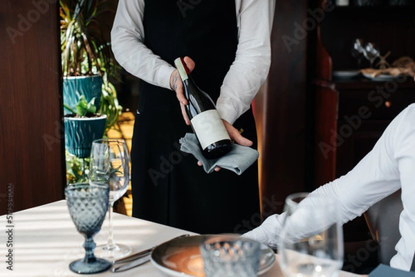 Fototapeta A young sommelier in a stylish uniform demonstrates and offers the client fine wine in the restaurant. Customer service. Table setting in a fine restaurant.