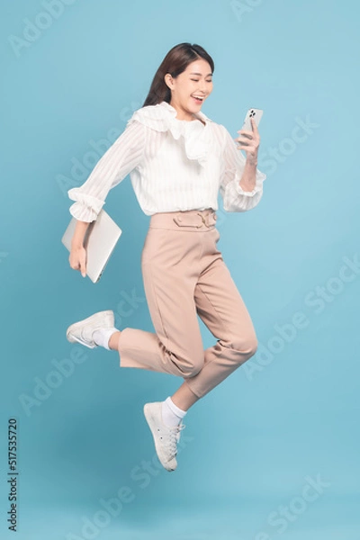 Fototapeta Young beautiful asian woman with smart casual cloth use smartphone and jump isolated on blue background