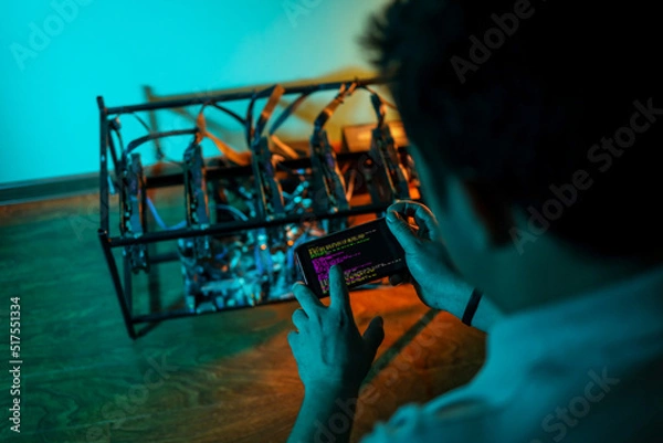 Fototapeta Male programmer adjusting cryptocurrency mining rig to optimal settings with mobile phone