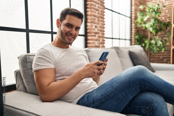 Fototapeta Young hispanic man using smartphone sitting on sofa at home