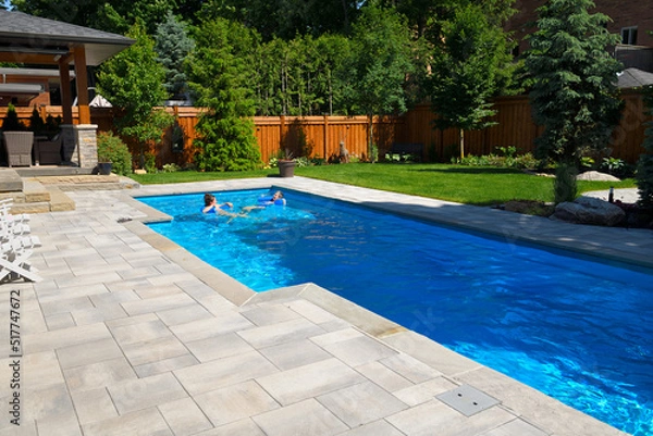 Fototapeta Mother and daughter swimming in new back yard pool with  patio of pavers and green lawn and gardens