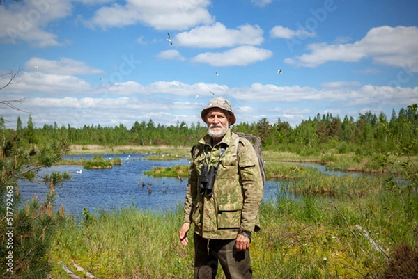 Obraz A man with a beard in camouflage clothes, with binoculars, an explorer, a tourist. Stands against the background of a forest lake, forest and blue sky with clouds.