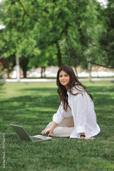 Fototapeta Beautiful Caucasian girl working with laptop computer sitting on green grass in summer park. Happy female outdoor working with technology, education and surfing the net, preparing for exams concept