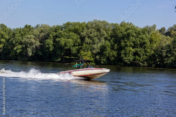 Fototapeta Modern speedboat sailing on forest background. Summer leisure and water activity. Rest On The River By Boat. Beautiful Motor Boat Rushes On The Water Surface Leaving Behind A Foam Trail