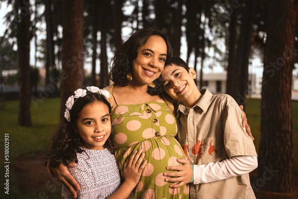 Obraz pregnant latin mom hugging her son and daughter in pine forest looking at the camera