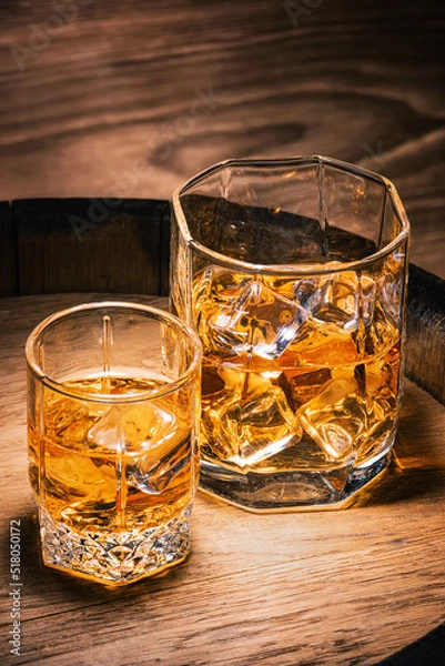 Fototapeta Brandy and whiskey in glasses with ice close-up on wooden barrel, vertical format. Whiskey and cognac in stacks of ice stand on wooden barrel against background of wooden wall.
