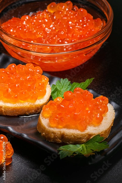 Fototapeta Red caviar in glass plate and on black plate on butter buns decorated with parsley on black background. Red caviar in cup and on butter rolls.