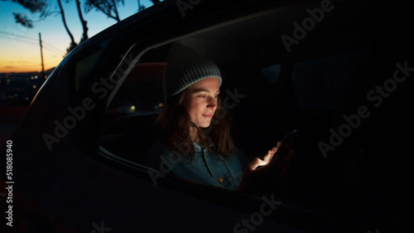 Obraz Side night cinematic view of young beautiful woman sit in taxi car back seat near window on road trip, hold, check on mobile phone, use smartphone, scroll social media news, read messages and smile
