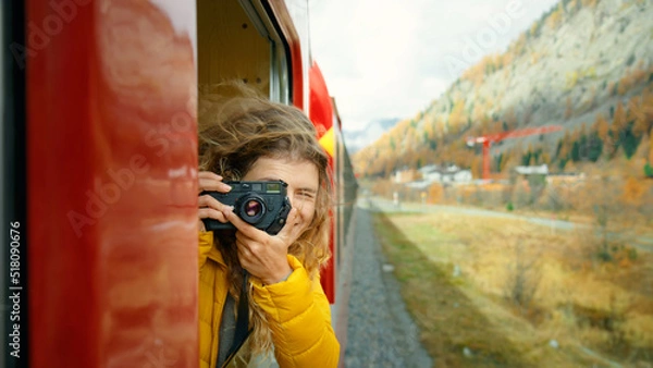 Fototapeta Inspiring female traveler and travel blogger look out of window of tourist train in cinematic beautiful mountains. Young beautiful woman hang out of window of red train. Excited world explorer