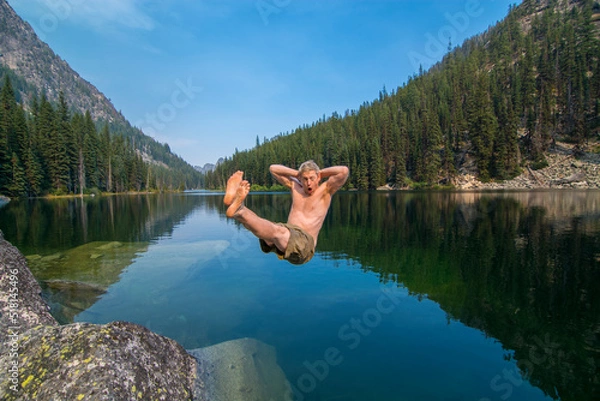 Fototapeta An adventurous athletic male hiker jumping backwards, with his hands behind his head in a lounging posture, into an alpine lake in Washington State.