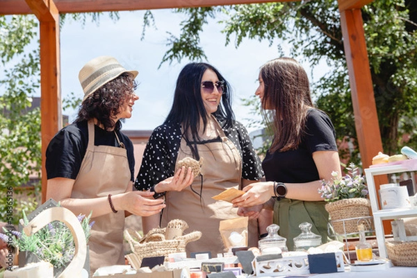 Fototapeta Three brunette women sellers of European appearance in aprons at a street fair laughing discuss their goods