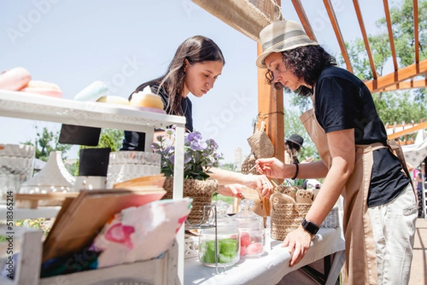 Fototapeta Young brunette girl buyer with long hair and woman seller in apron and hat on either side of the counter with goods at the street fair in summer