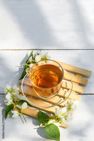 Obraz Jasmine tea in glass cup and flowers on a wooden white background, natural light photo with hard shadows. Cup with green jasmin tea