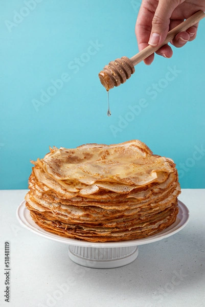 Fototapeta Stack of French crepes or Russian blini, thin pancakes on white plate on blue, turquoise background. Woman's hand pours honey on pancakes. Honey drips from wooden spoon on crepes or blini.
