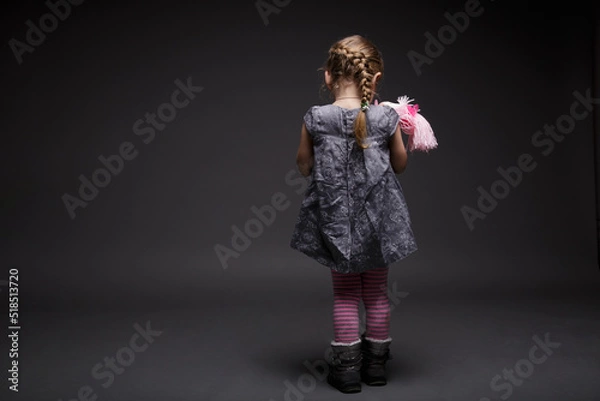 Fototapeta Rear image of a little girl with doll, toddler, offended by someone, being in bad mood, posing isolated over dark grey background.
