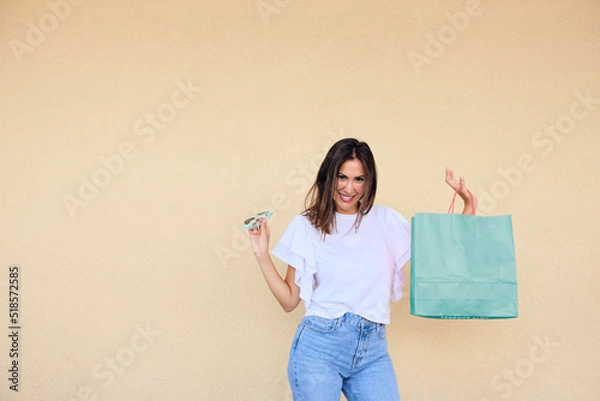 Obraz happy young woman enjoying a day of shopping with a bag in one hand and sunglasses in the other.