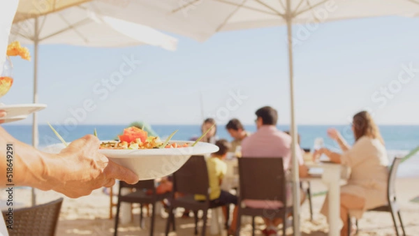 Fototapeta Professional waiter bringing different plates - paella, salad, prawns in tempura - to a family's table in a restaurant on the beach