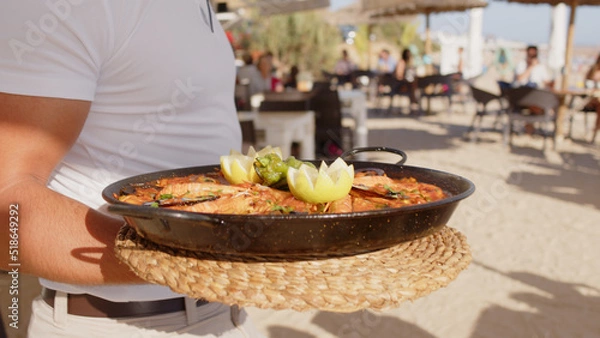 Fototapeta Professional waiter bringing different plates - paella, salad, prawns in tempura - to a family's table in a restaurant on the beach