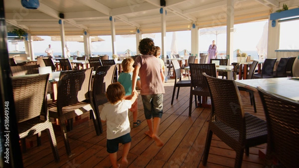 Fototapeta 4 sibling boys look at the camera smiling, play while running through a restaurant in front of the camera, eat ice cream, hug their mother
