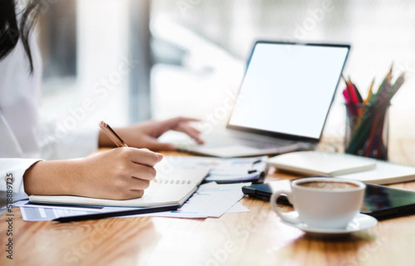 Fototapeta Close up view, young asian businesswoman sitting on desk in office writing her plan on note book, use computer laptop working with paperwork of investment, select focus on hand holding pen