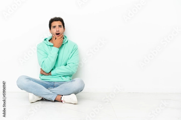 Fototapeta Caucasian handsome man sitting on the floor surprised and shocked while looking right