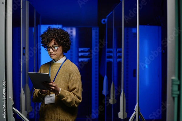 Fototapeta Young female engineer typing data in digital tablet while standing near server racks