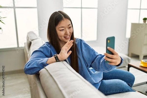 Obraz Young chinese girl having video. call using smartphone sitting on the sofa at home.