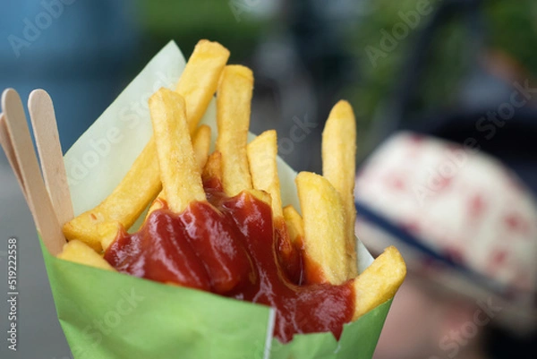 Fototapeta Close up a portion of fries with ketchup in a paper cone with some wooden forks. Fried street food outside, a blurred child's hat in the background.