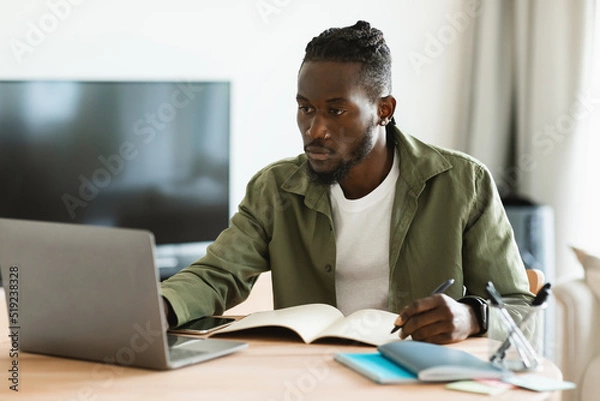 Fototapeta Focused african american man using laptop, working remotely and taking notes, having remote meeting from home
