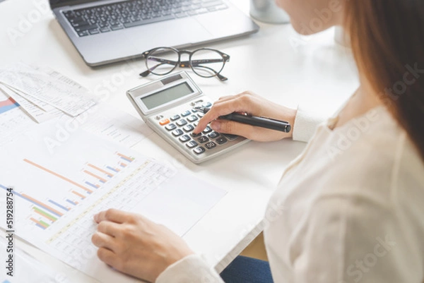 Fototapeta Account finance, asian young business woman hand use calculator for calculate budget, cost and income of company from charts, reports paperwork, plan spend money expenses, working on desk at home.