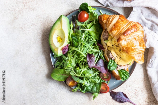 Obraz Croissant with tuna, poached egg, avocado and hollandaise sauce served with green salad leaves and tomatoes. Croissant sandwich with egg benedict and tuna.