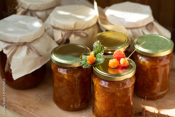 Fototapeta on a wooden and fabric background there are beautiful glass jars with orange sweet jam from orange berries of northern cloudberries from Karelia. several fresh berries lie on golden lids with a green 