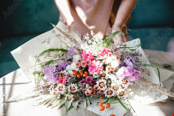 Fototapeta A woman florist packs a multi-branch bouquet in white paper on the table in her flower studio. Only the bouquet in the frame