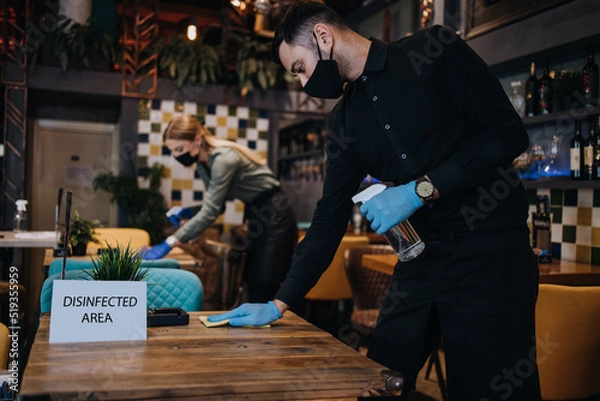 Fototapeta Young restaurant waiters cleaning and disinfecting tables and surfaces during Coronavirus pandemic disease. They are wearing protective face masks and gloves.