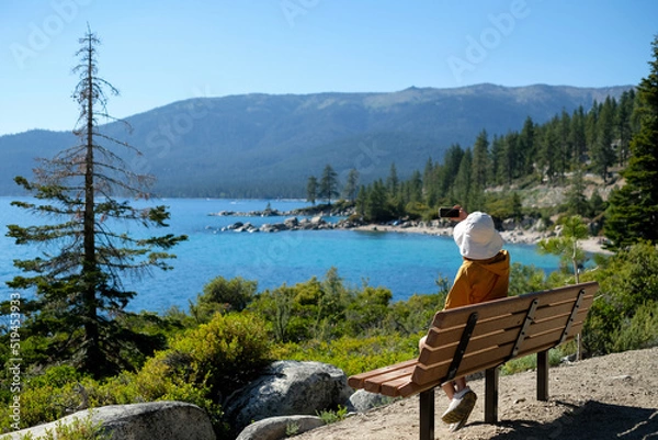 Fototapeta Woman is taking a picture of lake Tahoe sitting on the bench.Traveling and exploring concept