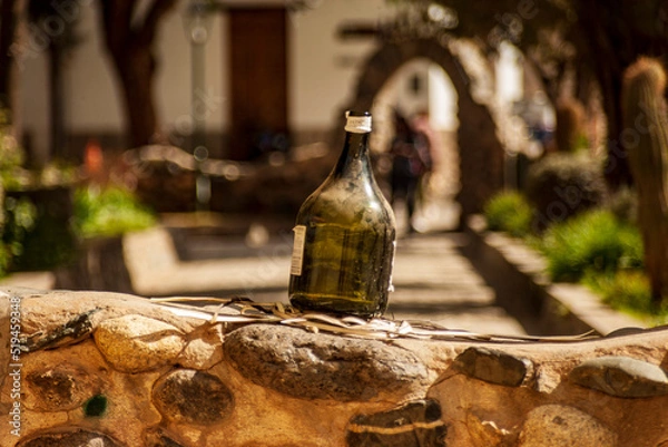Obraz Horizontal format photograph of a bottle of green wine without content on a small white wall in a square in the town of Cachi, Salta, Argentina.