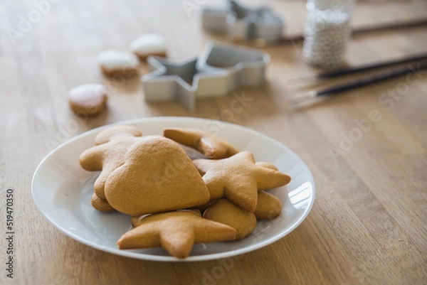 Fototapeta A white plate of traditional ginger biscuits stands on a wooden table. Brushes for drawing on gingerbread, forms for gingerbread in the form of hearts and stars.